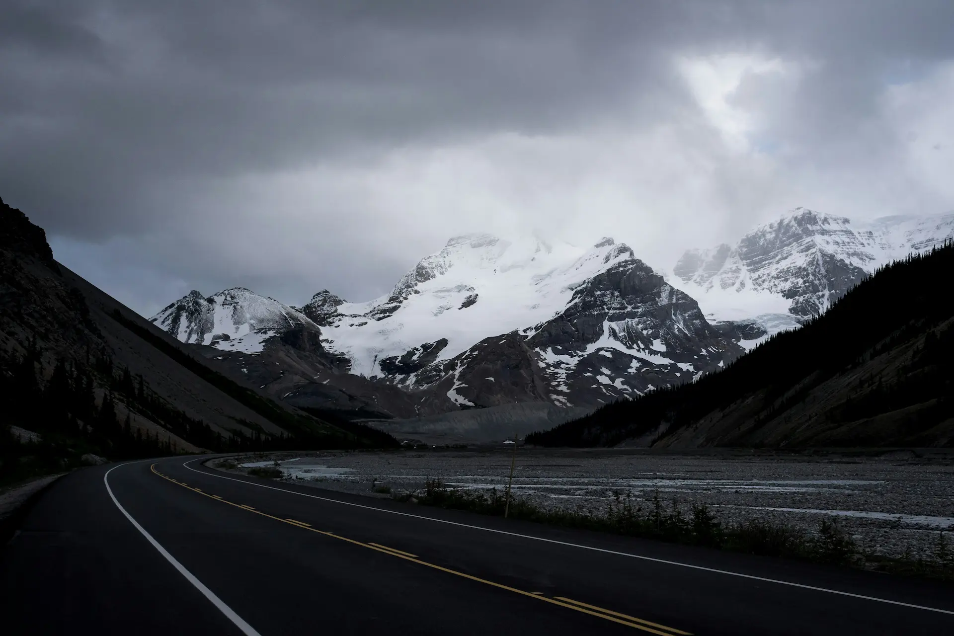 Curved highway with yellow centre line leading toward snow-capped mountain peaks, forested valleys, and a glacial lake under overcast sky