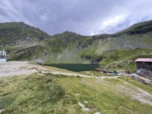 Alpine lake nestled in a mountain valley with steep rocky slopes, green meadows, and a small cabin with a pink roof on the right side
