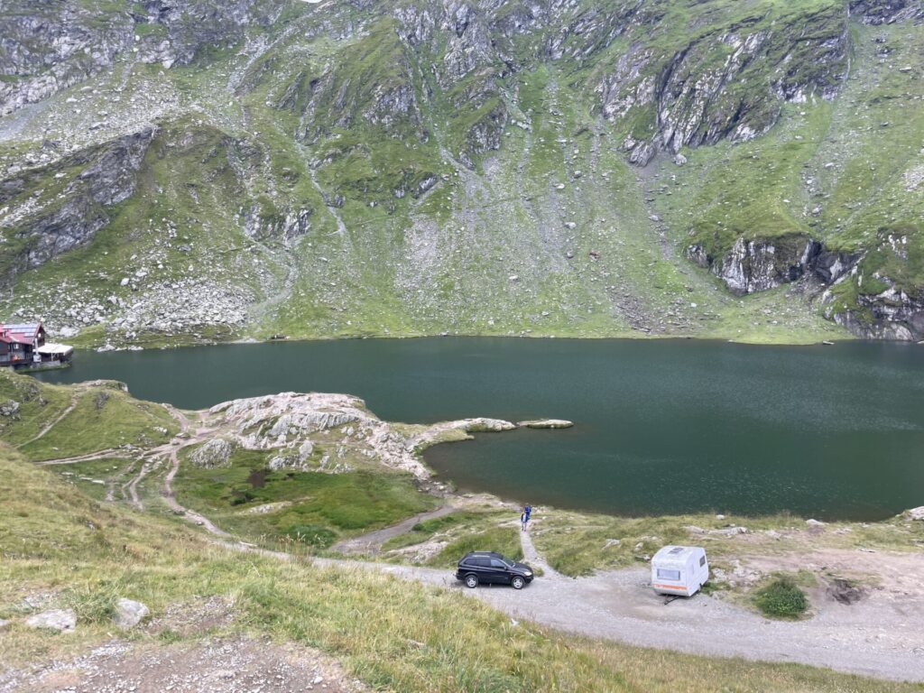 Alpine lake surrounded by steep rocky slopes with green meadows, a parked car and caravan on a gravel road, and a small red building near the water's edge