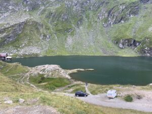 Alpine lake surrounded by steep rocky slopes with green meadows, a parked car and caravan on a gravel road, and a small red building near the water's edge