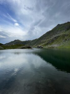 Alpine lake surrounded by steep green and rocky mountains, with a small cabin and buildings visible on the far shore under a partly cloudy sky
