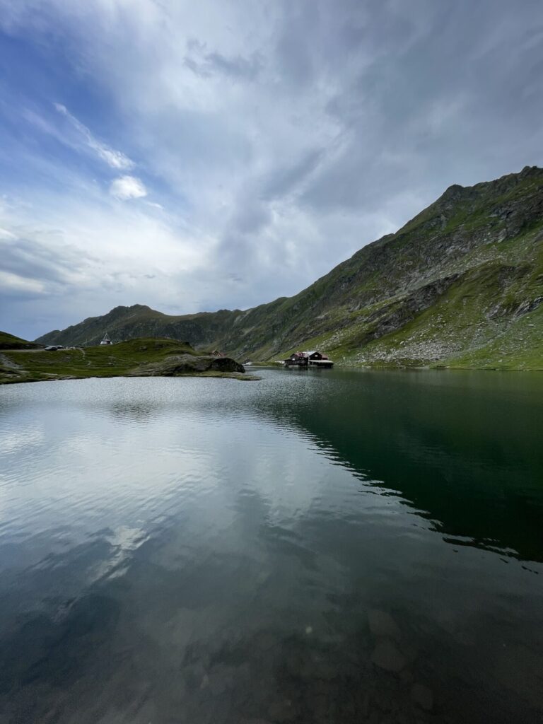 Alpine lake surrounded by steep green and rocky mountains, with a small cabin and buildings visible on the far shore under a partly cloudy sky