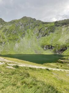 Alpine lake nestled beneath steep green mountains with exposed grey rock outcrops and grassy meadow in foreground