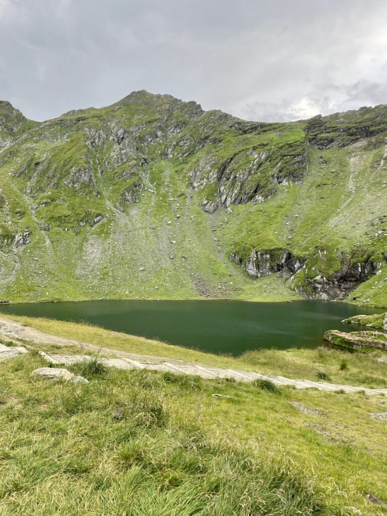 Alpine lake nestled beneath steep green mountains with exposed grey rock outcrops and grassy meadow in foreground