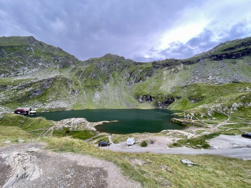 Alpine lake surrounded by rocky green mountains, with a small cabin and parked vehicles at the shore under a partly cloudy sky