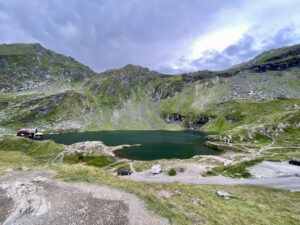 Alpine lake surrounded by rocky green mountains, with a small cabin and parked vehicles at the shore under a partly cloudy sky
