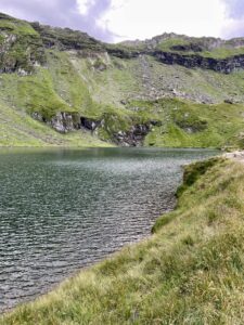 Alpine lake surrounded by steep green hillsides with exposed rock outcrops and grassy foreground under cloudy sky
