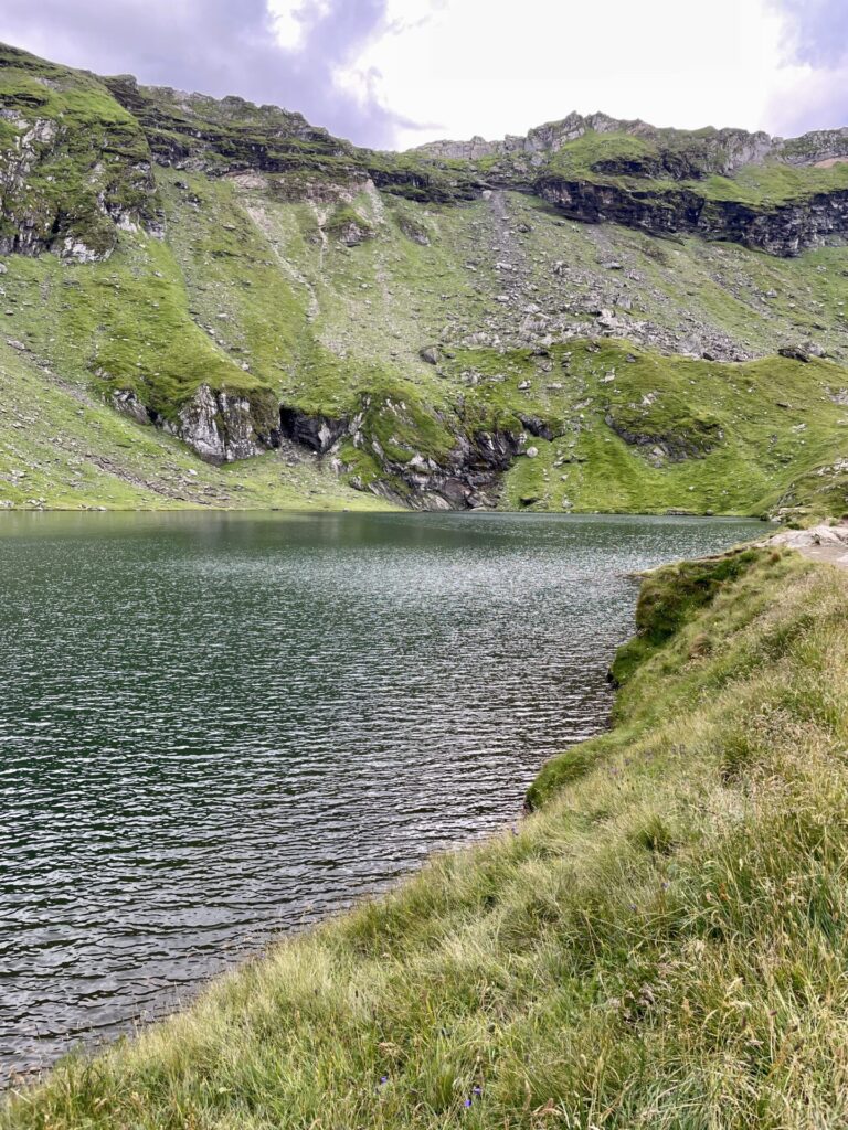 Alpine lake surrounded by steep green hillsides with exposed rock outcrops and grassy foreground under cloudy sky
