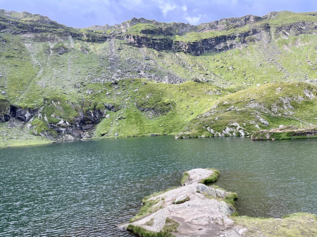 Alpine lake surrounded by steep green hillsides with exposed rock outcrops and layered cliff faces under blue sky