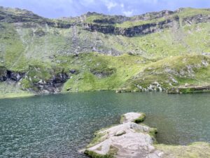 Alpine lake surrounded by steep green hillsides with exposed rock outcrops and layered cliff faces under blue sky