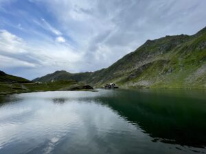 Alpine lake surrounded by steep green mountains with scattered buildings on the far shore under a partly cloudy sky