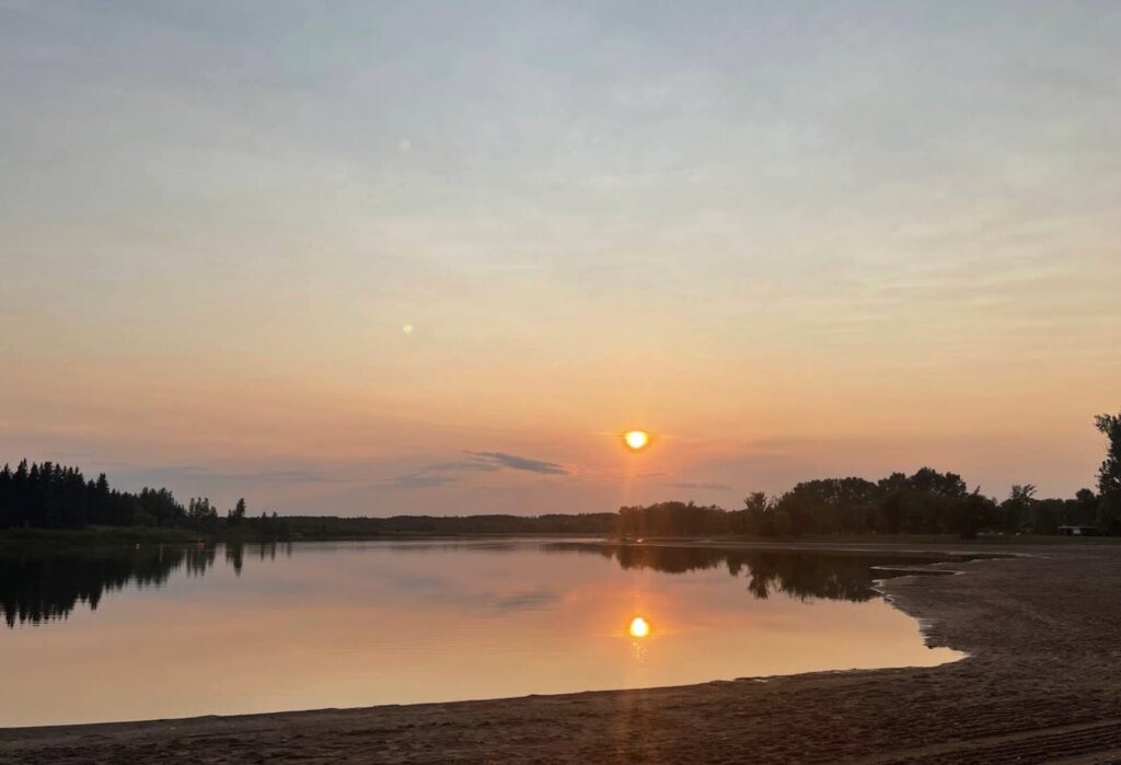 Sunset over a calm river with sandy beach in foreground, forested shoreline, and sun reflecting on water