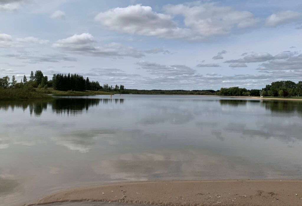 Calm lake with sandy beach in foreground, forested shoreline and partly cloudy sky reflected in still water