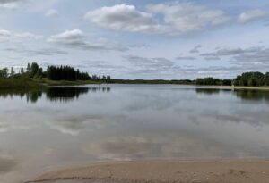 Calm lake with sandy beach in foreground, forested shoreline and partly cloudy sky reflected in still water