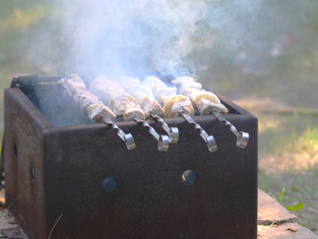 Meat skewers cooking over a campfire in a metal grill box, with smoke rising from the food