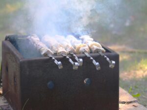 Meat skewers cooking over a campfire in a metal grill box, with smoke rising from the food