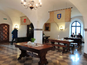 Interior of a medieval castle hall with wooden tables, heraldic banners displaying red and blue shields with crosses, arched ceilings, a chandelier, and visitors exploring the space