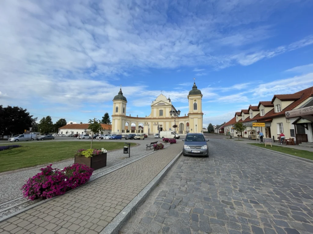 Town square with a cream-coloured church featuring two green domes and crosses, flanked by residential buildings with red-tiled roofs, paved plaza with magenta flowers, parked cars, and blue sky with wispy clouds