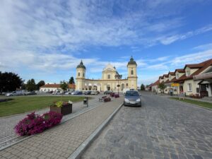 A cream-coloured church with two green-domed towers and a cross stands in a town square, flanked by parked cars and residential buildings with red-tiled roofs under a blue sky with wispy clouds