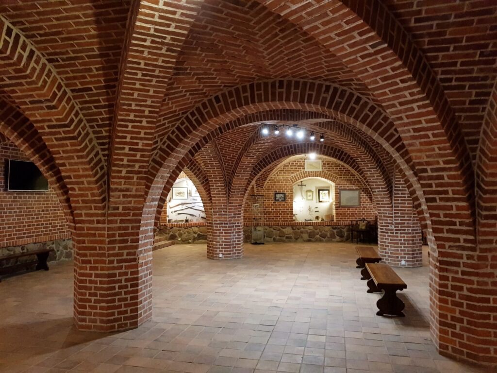 Brick vaulted underground chamber with arched ceilings, supporting columns, and benches, lit by overhead lights and leading to a doorway at the far end