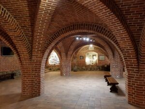 Brick vaulted underground chamber with arched ceilings, supporting columns, and benches, lit by overhead lights and leading to a doorway at the far end