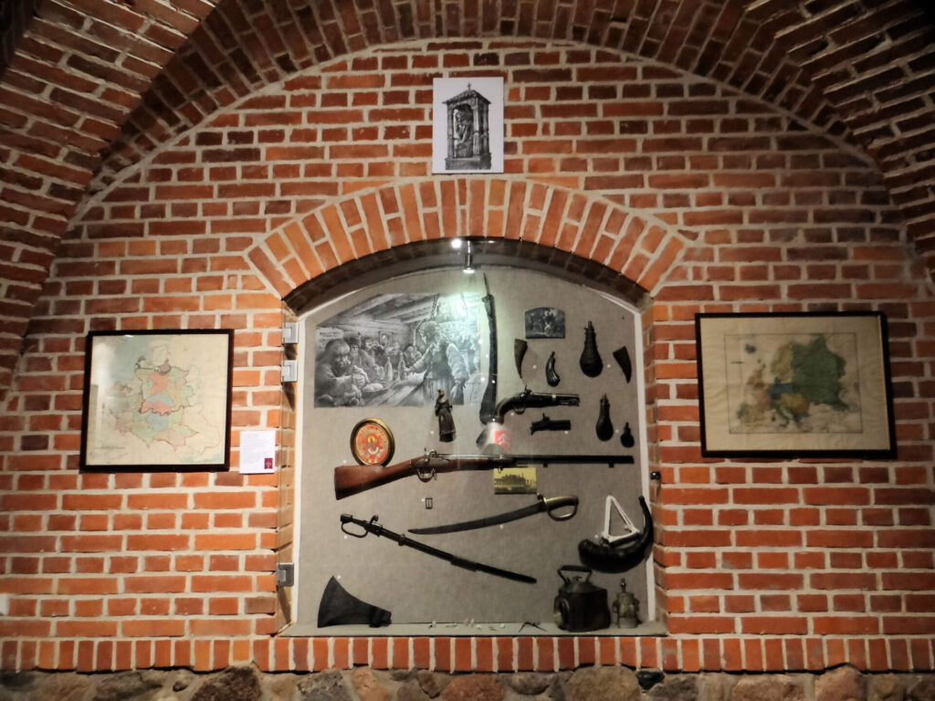 Museum display case built into a brick archway, containing historical military artifacts including a rifle, powder horns, swords, and period maps on the surrounding walls