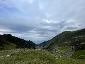Alpine valley with winding mountain road, steep green slopes, rocky outcrops, and distant lowlands under cloudy sky