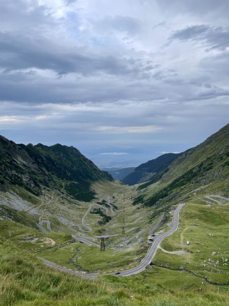 Winding mountain road with tight hairpin curves descending through a lush green valley between steep forested peaks under a cloudy sky