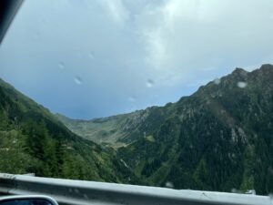 Alpine valley with forested slopes and rocky peaks under blue sky, viewed from a vehicle