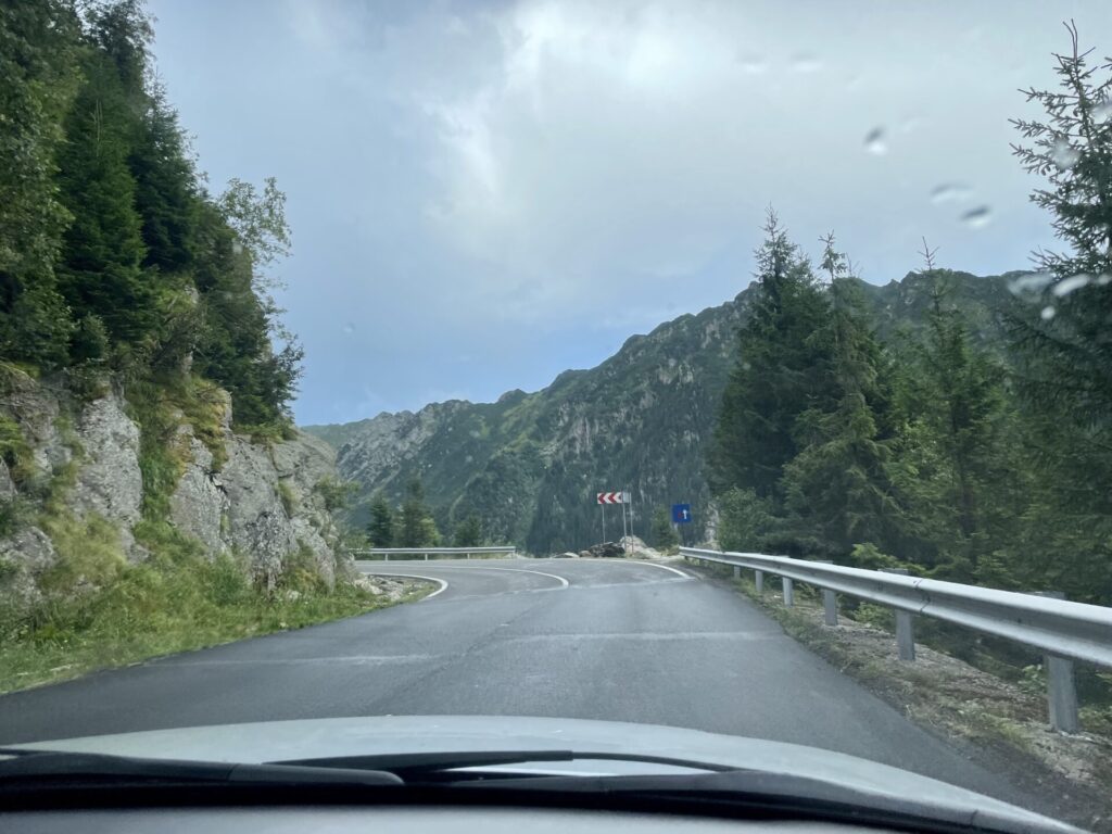 Mountain road curving through a forested valley with rocky cliffs, guard rails, and directional signage visible ahead
