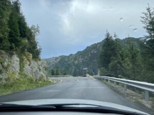 Mountain road curving through a forested valley with rocky cliffs, guard rails, and directional signage visible ahead