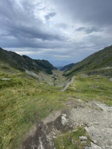 Winding mountain road through a green alpine valley with steep rocky slopes on either side and dramatic cloudy sky above