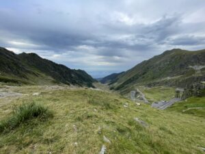 Alpine mountain valley with winding road through grassy slopes and rocky peaks under cloudy sky