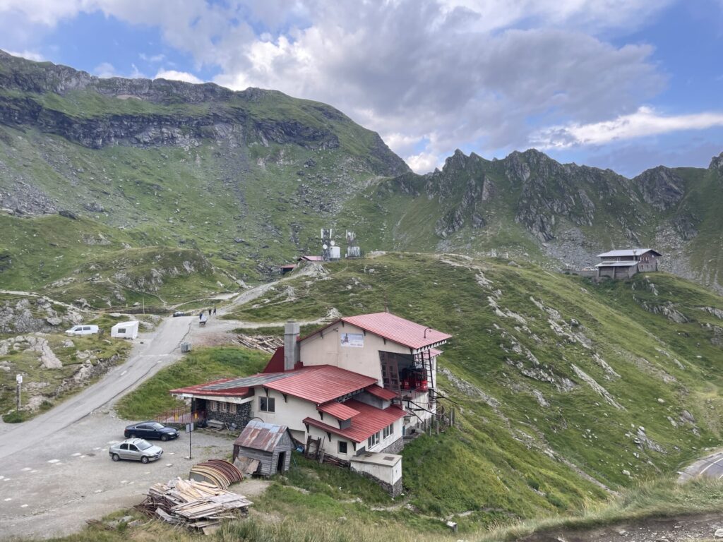 Mountain valley settlement with a multi-storey building featuring red metal roofing, parked vehicles, and rocky peaks with green slopes in the background