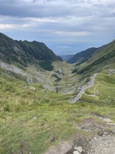 Winding mountain road with multiple hairpin turns descending through a green alpine valley between steep rocky slopes, with distant plains visible beyond the valley