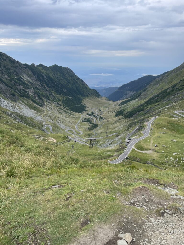 Winding mountain road with multiple hairpin turns descending through a green alpine valley between steep rocky slopes, with distant plains visible beyond the valley