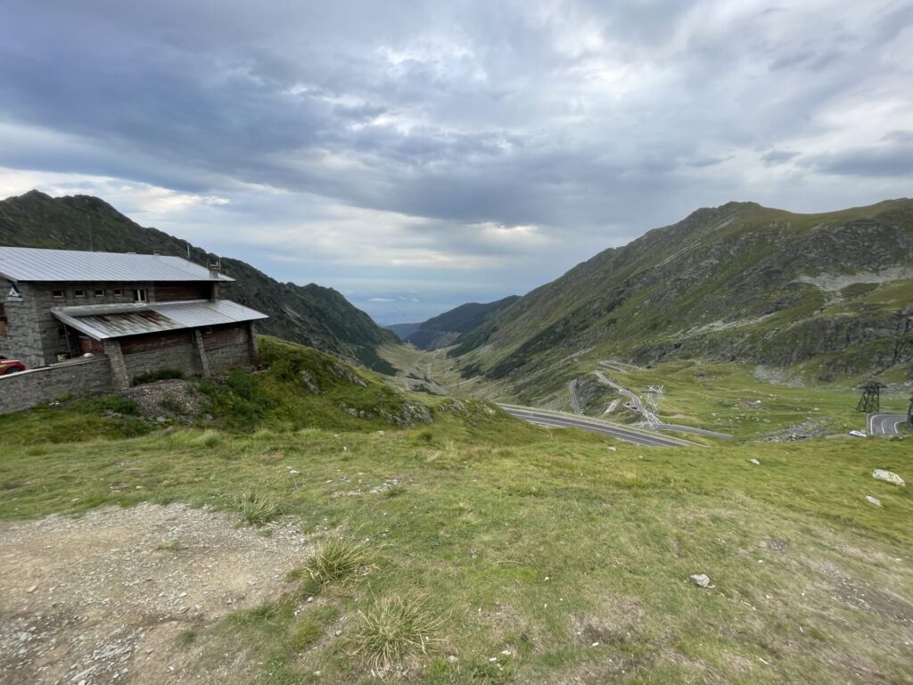 Mountain refuge with metal roof beside a winding road through a green alpine valley under cloudy skies