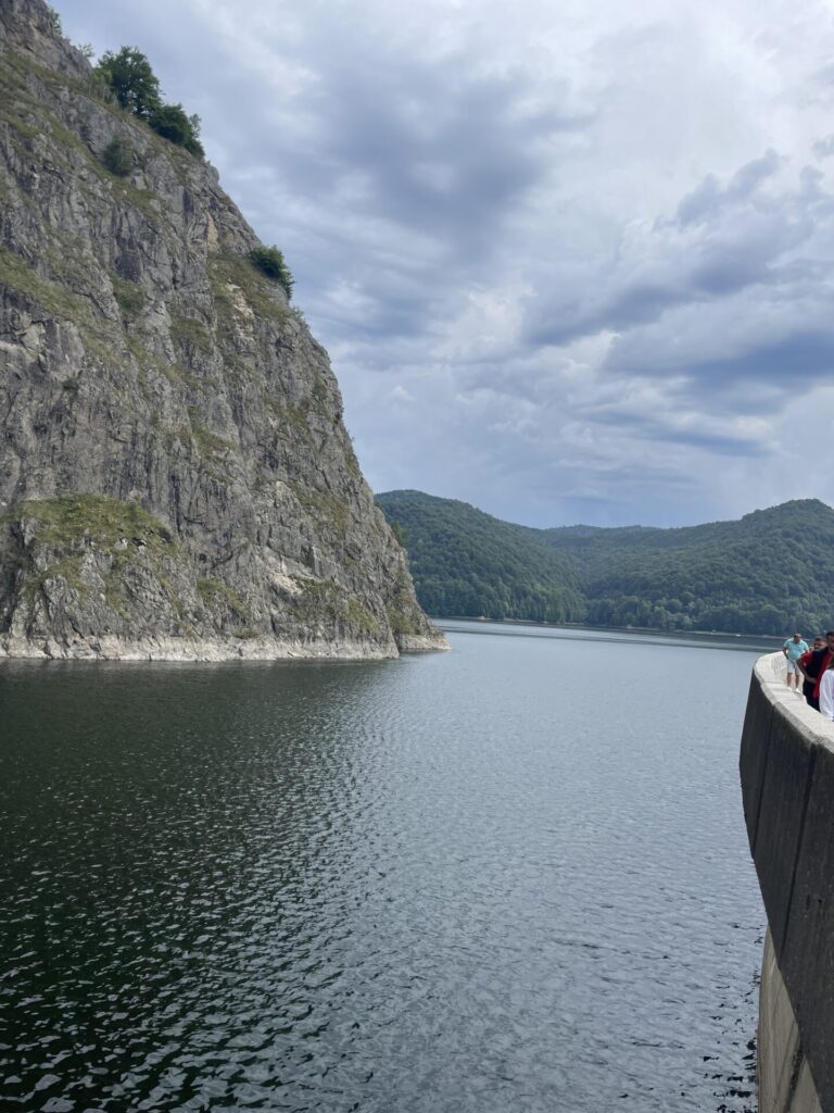 Steep limestone cliff with vegetation rising above calm lake waters, forested hills visible across the water, taken from a boat