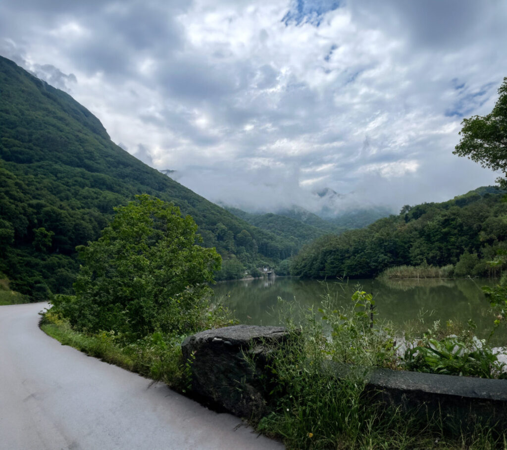Scenic mountain valley with a calm river, lush green forested slopes on both sides, and mist-shrouded peaks under a cloudy sky