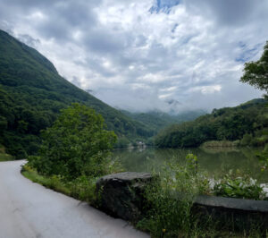 Scenic mountain valley with a calm river, lush green forested slopes on both sides, and mist-shrouded peaks under a cloudy sky