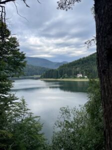 A calm lake surrounded by dense coniferous forest and mountains under a cloudy sky, with a small building visible on the far shoreline