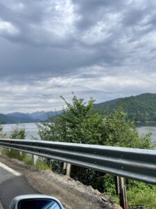 Mountain lake surrounded by forested hills under cloudy sky, viewed from a roadside with metal guardrail in foreground