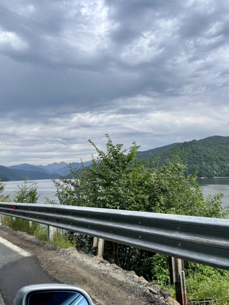 Mountain lake surrounded by forested hills under cloudy sky, viewed from a roadside with metal guardrail in foreground