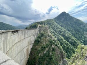 Concrete dam spanning a forested valley with steep, densely vegetated mountains and a communication tower visible on the distant ridge