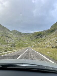 Straight road stretching through a mountain valley with green slopes, rocky outcrops, and scattered buildings under a cloudy sky, photographed from inside a vehicle