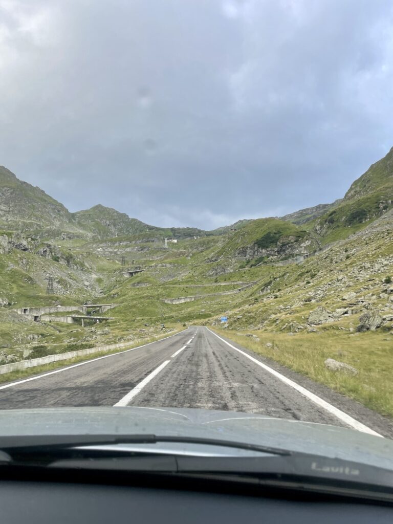 Straight road stretching through a mountain valley with green slopes, rocky outcrops, and scattered buildings under a cloudy sky, photographed from inside a vehicle