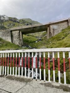 Concrete bridge spanning a mountain stream with red and white safety railings in the foreground, surrounded by green hillsides and rocky terrain