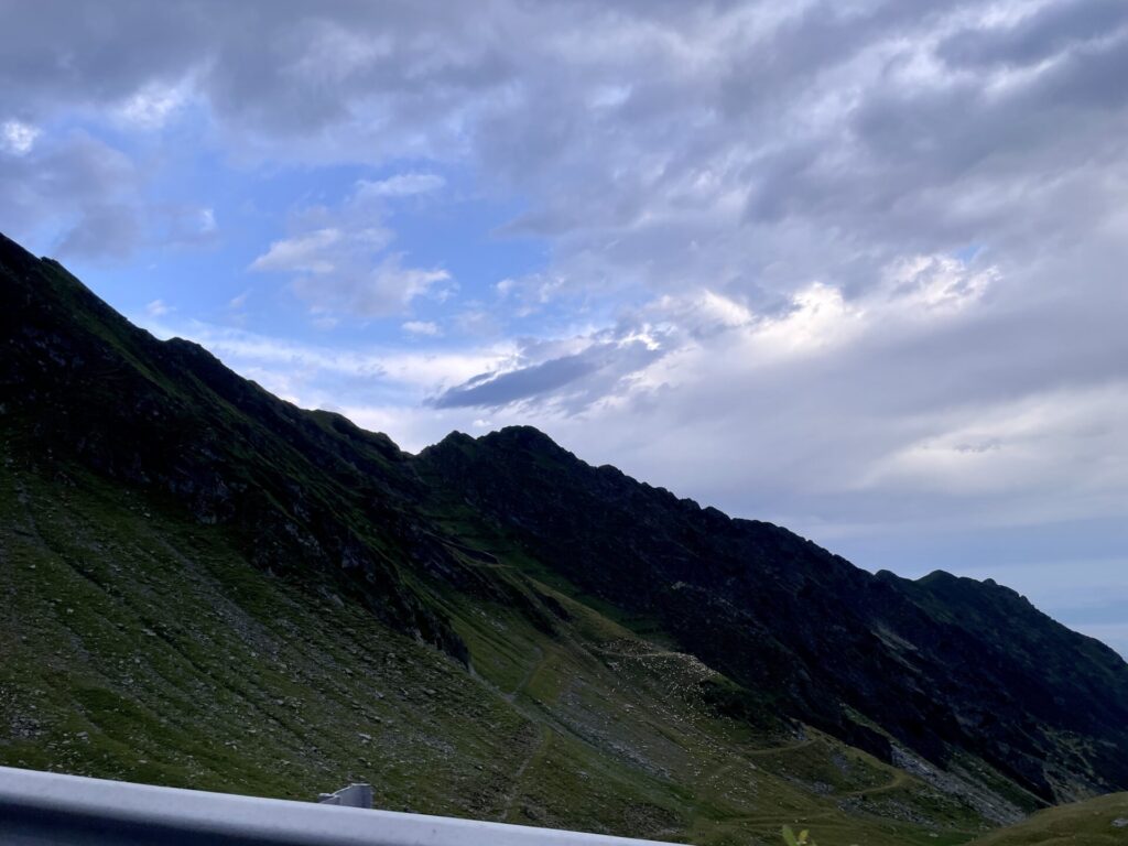 Alpine valley with snow-capped mountain peak in the distance, green slopes in foreground, and dramatic cloudy sky