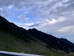 Alpine valley with snow-capped mountain peak in the distance, green slopes in foreground, and dramatic cloudy sky