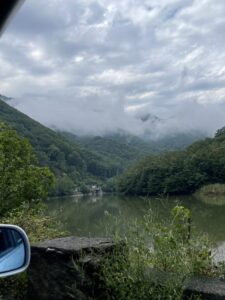 Scenic river valley with forested mountains shrouded in mist, taken from a vehicle viewpoint with a car mirror visible in the foreground